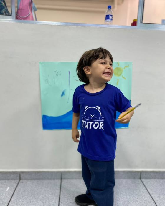 Menino sorrindo com o pincel na mão ao lado da sua pintura